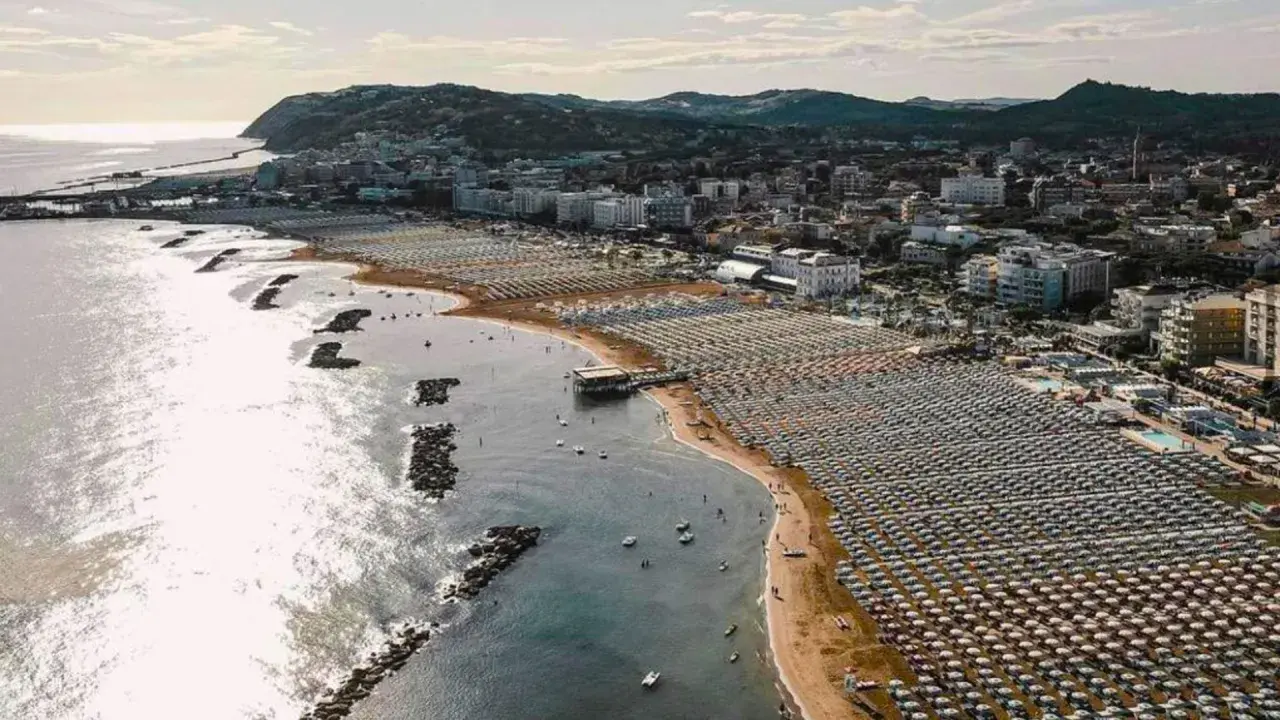 Vista aerea della cattolica spiaggia, con file ordinate di ombrelloni e lettini che si estendono lungo la costa, mentre il mare luccica sotto il sole.