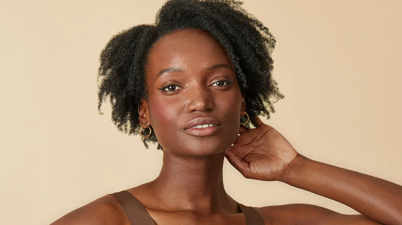 Une femme à la peau foncée, aux cheveux afro volumineux et bouclés, porte des boucles d'oreilles dorées.