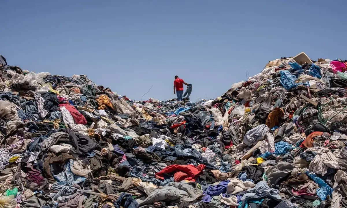 Un uomo in maglia rossa cammina su una montagna di vestiti scartati, simbolo dell'inquinamento tessile.