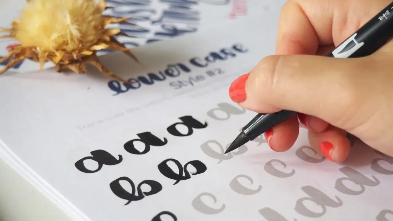Mano practicando cómo hacer lettering con un rotulador sobre un papel con letras de ejemplo.