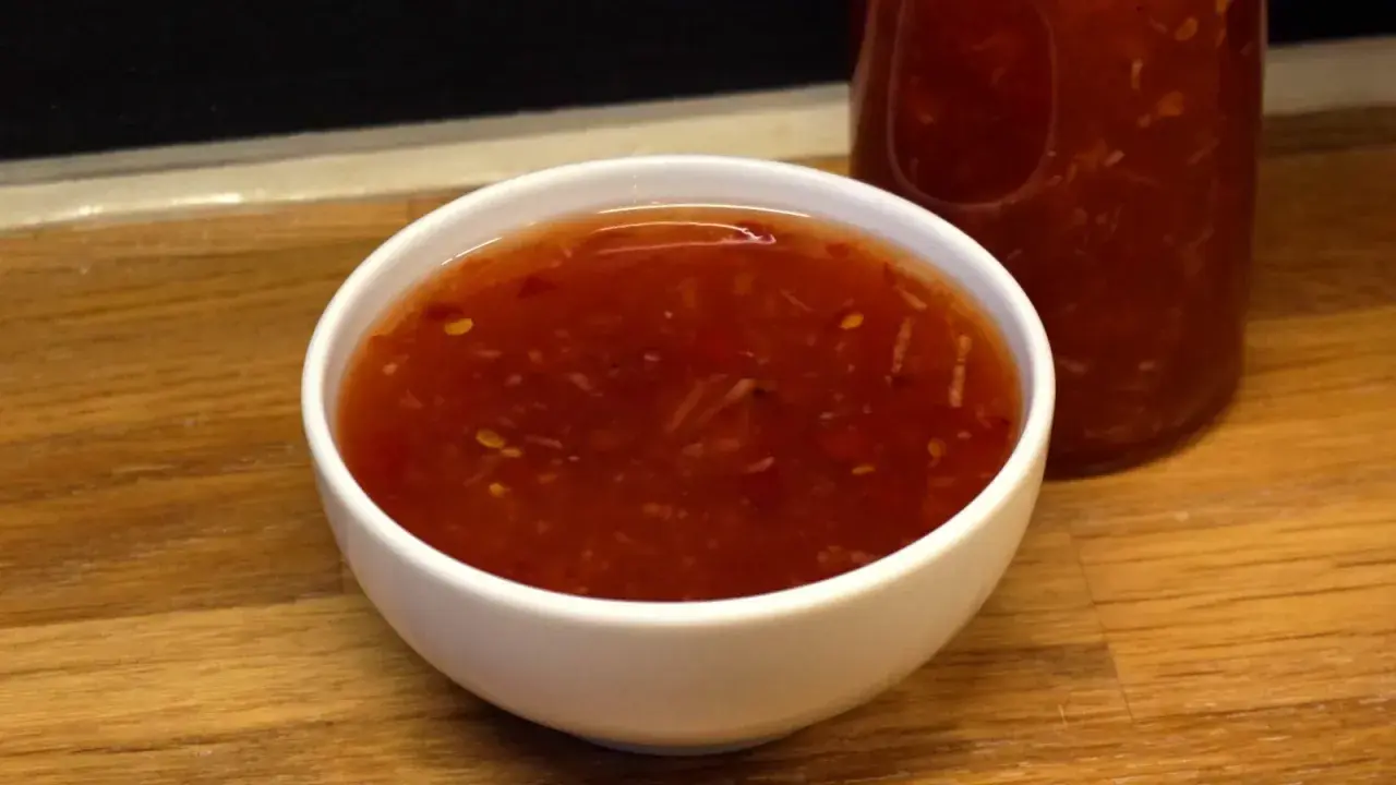 En skål med glansig, röd sweet chili sås med små bitar av chili och vitlök. En flaska med samma sås syns i bakgrunden.