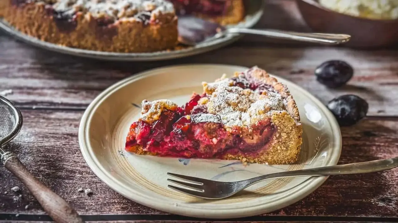Ein saftiger Zwetschgenkuchen mit Dinkelmehl vom Blech, bestäubt mit Puderzucker. Ein Stück auf einem Teller mit einer Gabel.