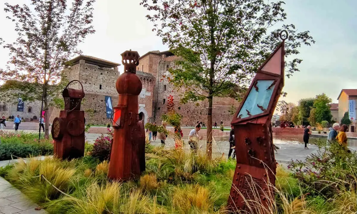 Sculture arrugginite e vegetazione rigogliosa nel Parco Fellini a Rimini, con il castello Sismondo sullo sfondo.