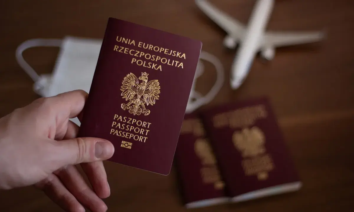 Dłoń trzyma polski paszport. W tle maseczka, samolot i inne paszporty.