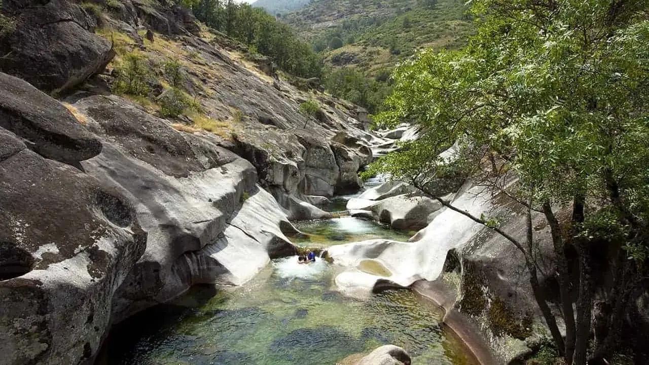 Un río de aguas cristalinas fluye entre rocas erosionadas, formando piscinas naturales. Dos personas disfrutan del baño en uno de los pilones, rodeados de vegetación y montañas.