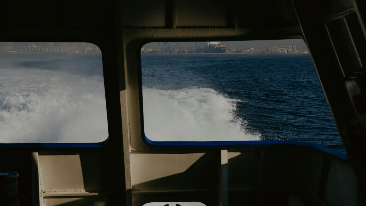 Vue depuis la cabine d'un bateau, avec le sillage blanc de l'eau. Au loin, une ville côtière et des grues de port. Les canaux VHF marine sont essentiels pour la communication.