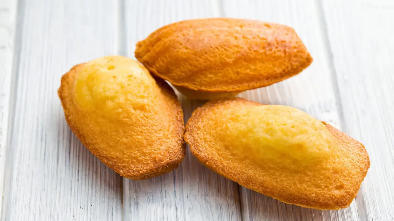 Trois madeleines dorées, tout juste sorties du four, sur une planche en bois. La cuisson madeleine est réussie, leur texture est parfaite.