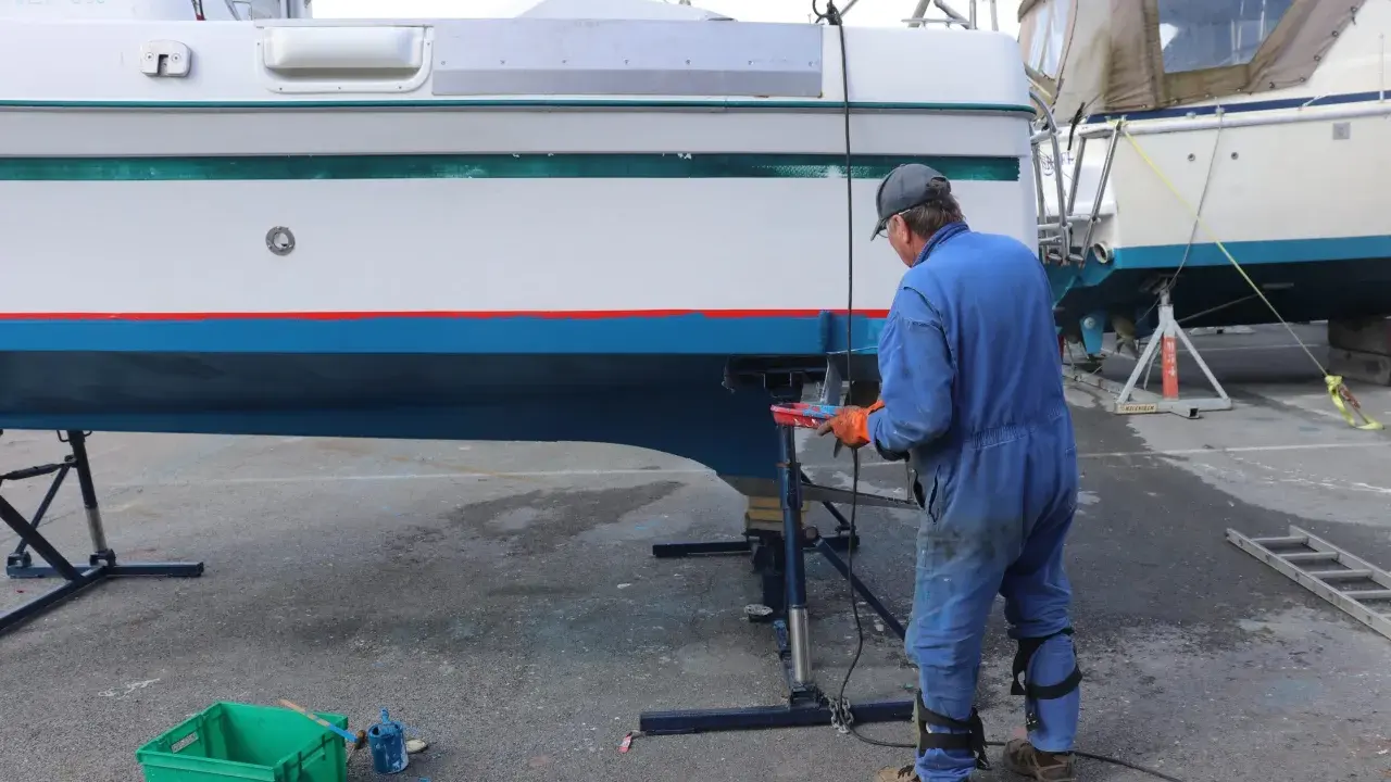 Un homme en combinaison bleue travaille sur la coque d'un bateau pour enlever l'antifouling. Des outils et des pots de peinture sont à proximité.