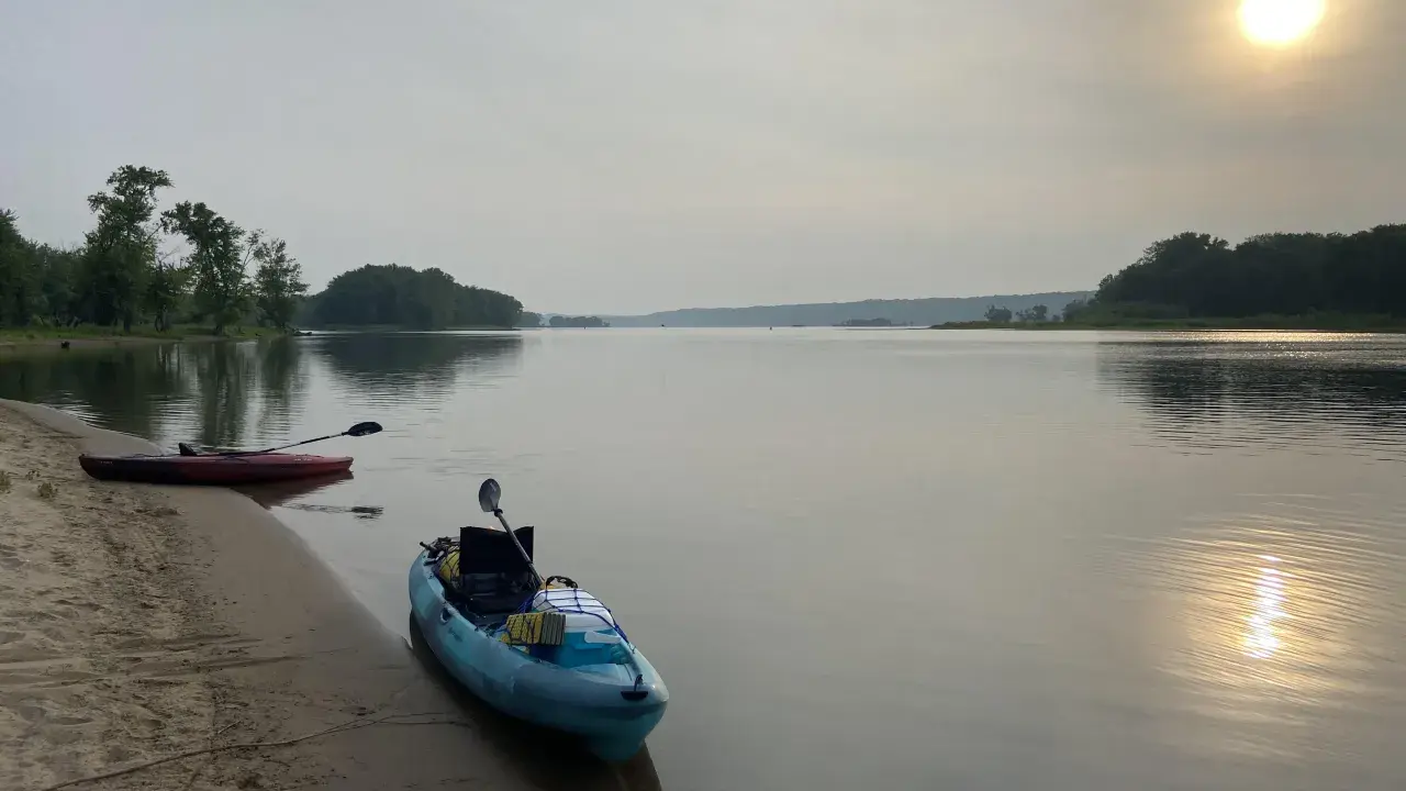 Två kajaker vid en sandstrand, redo för en paddling. Solen lyser svagt genom diset. Frågan är hur långt paddlar man på en dag?