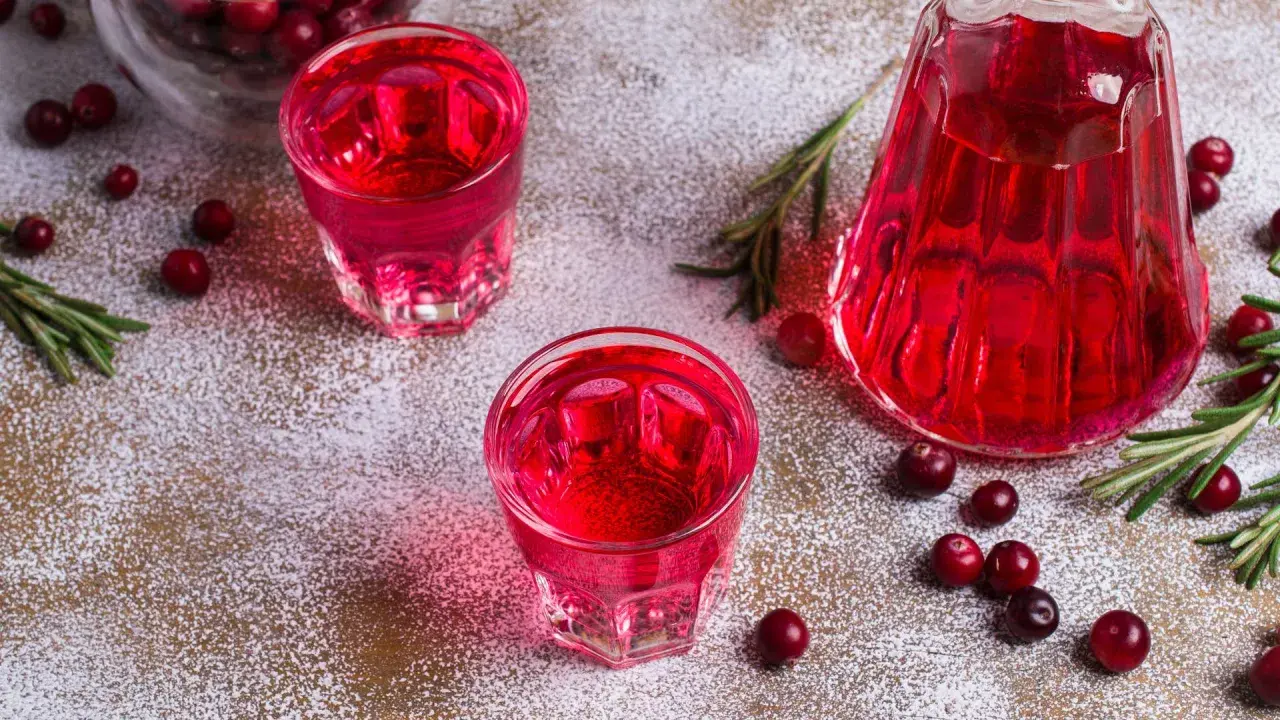 Två glas med röd vargtass drink, en karaff, tranbär och rosmarin på ett pudrat underlag.