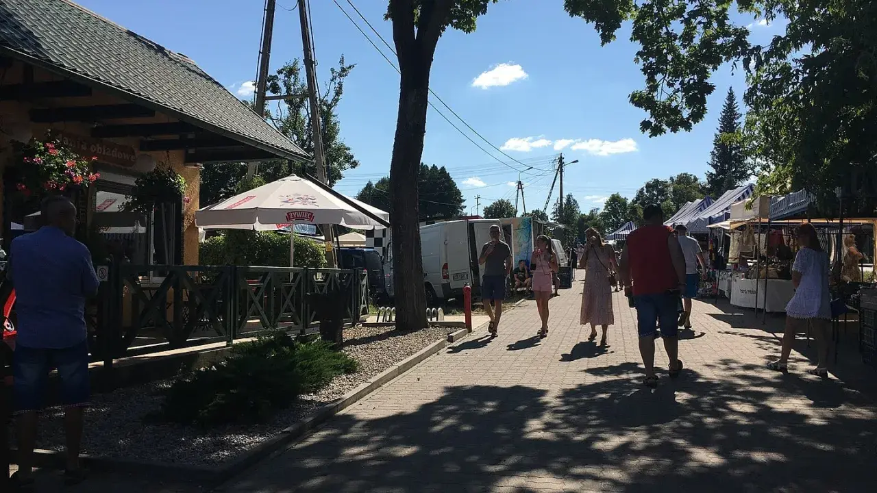 Ludzie spacerują po targu, podziwiając **tleń atrakcje**. Widać stoiska z towarami, restaurację i drzewa rzucające cień.