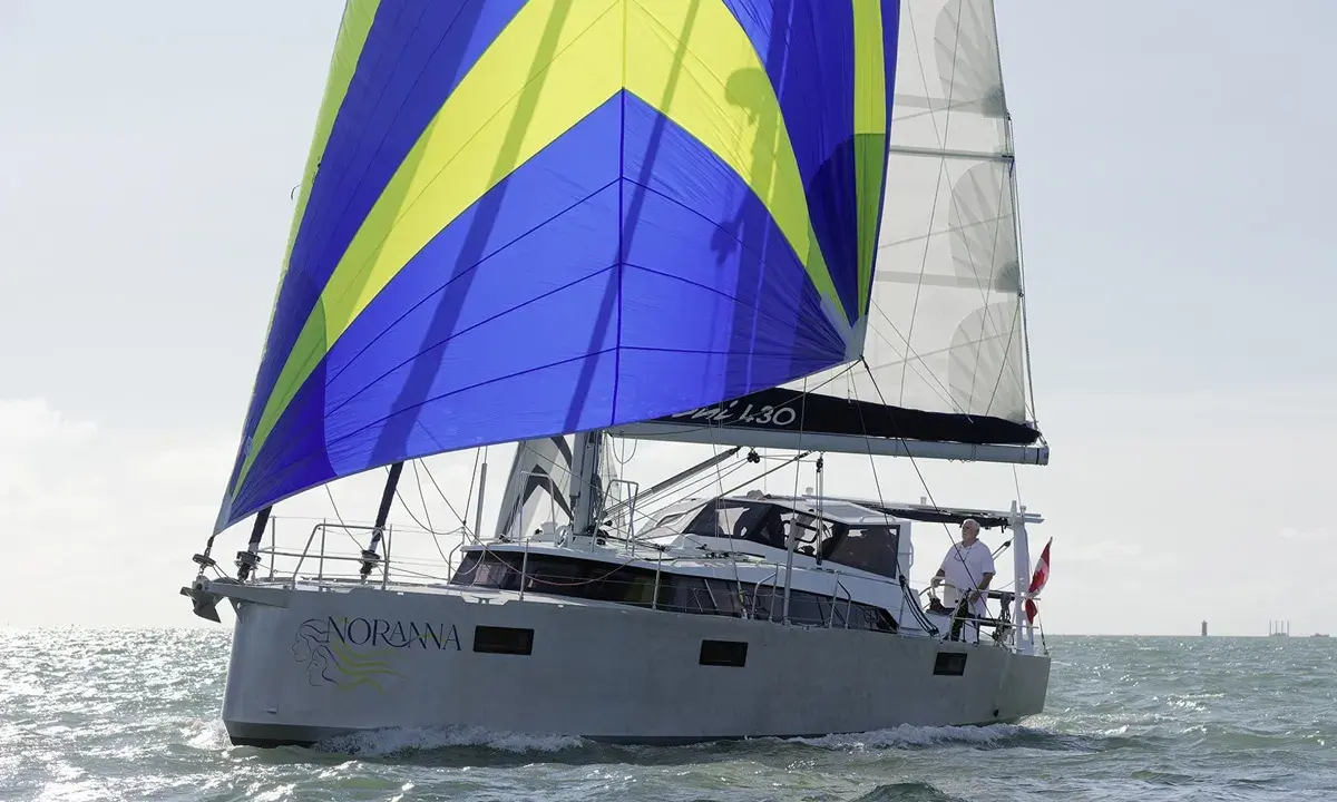 Le voilier "Noranna" L430 navigue sur une mer agitée, sa grand-voile aux couleurs vives bleu et jaune captant le vent.