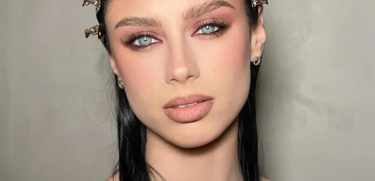 Ojos ahumados en tonos rosados y marrones, realzando unos ojos azules penetrantes. Labios nude y cabello oscuro completan el look.