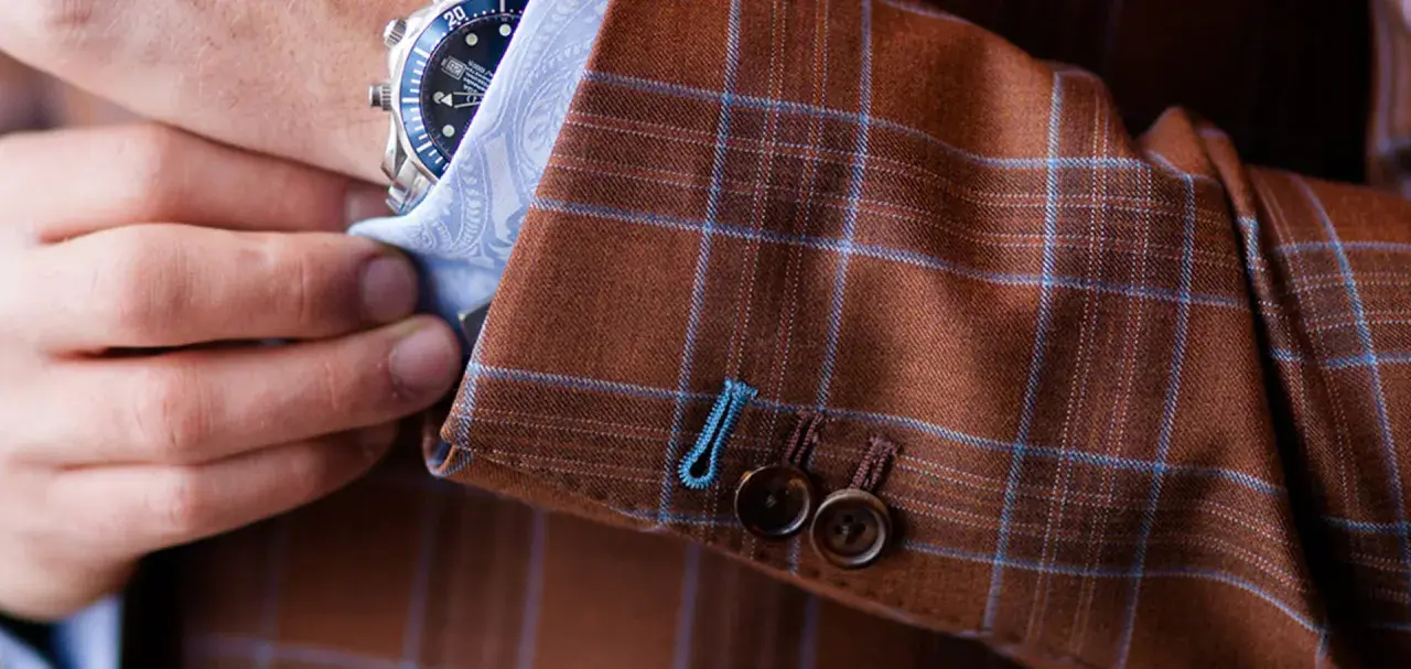 A man adjusts his watch on his wrist, revealing the detailed pick stitching on the cuff of his brown plaid suit jacket.