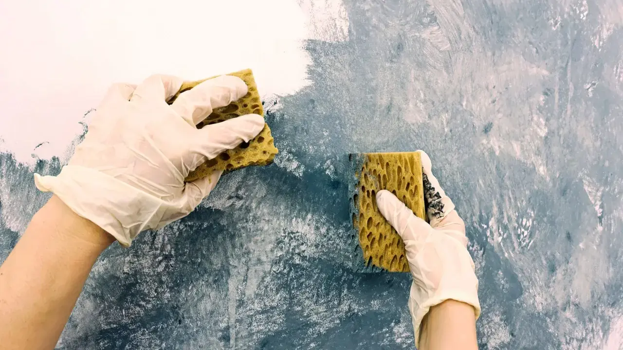 Händer med handskar målar akryl med svamp på en vägg för att skapa en texturerad effekt.