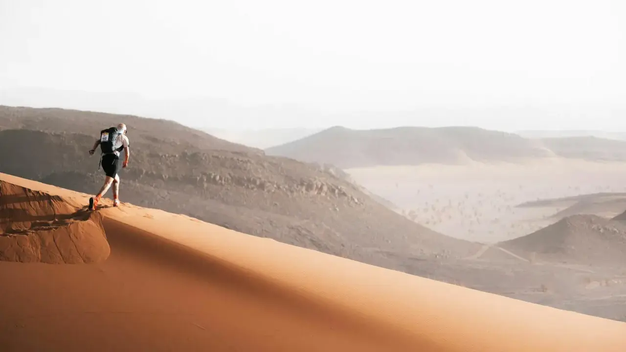 Läufer erklimmt eine Sanddüne, bereit für den Marathon des Sables. Die Wüstenlandschaft erstreckt sich in der Ferne.