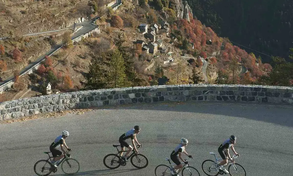 Ciclisti affrontano la salita del Col de la Couillole, con un villaggio arroccato sulla montagna autunnale.