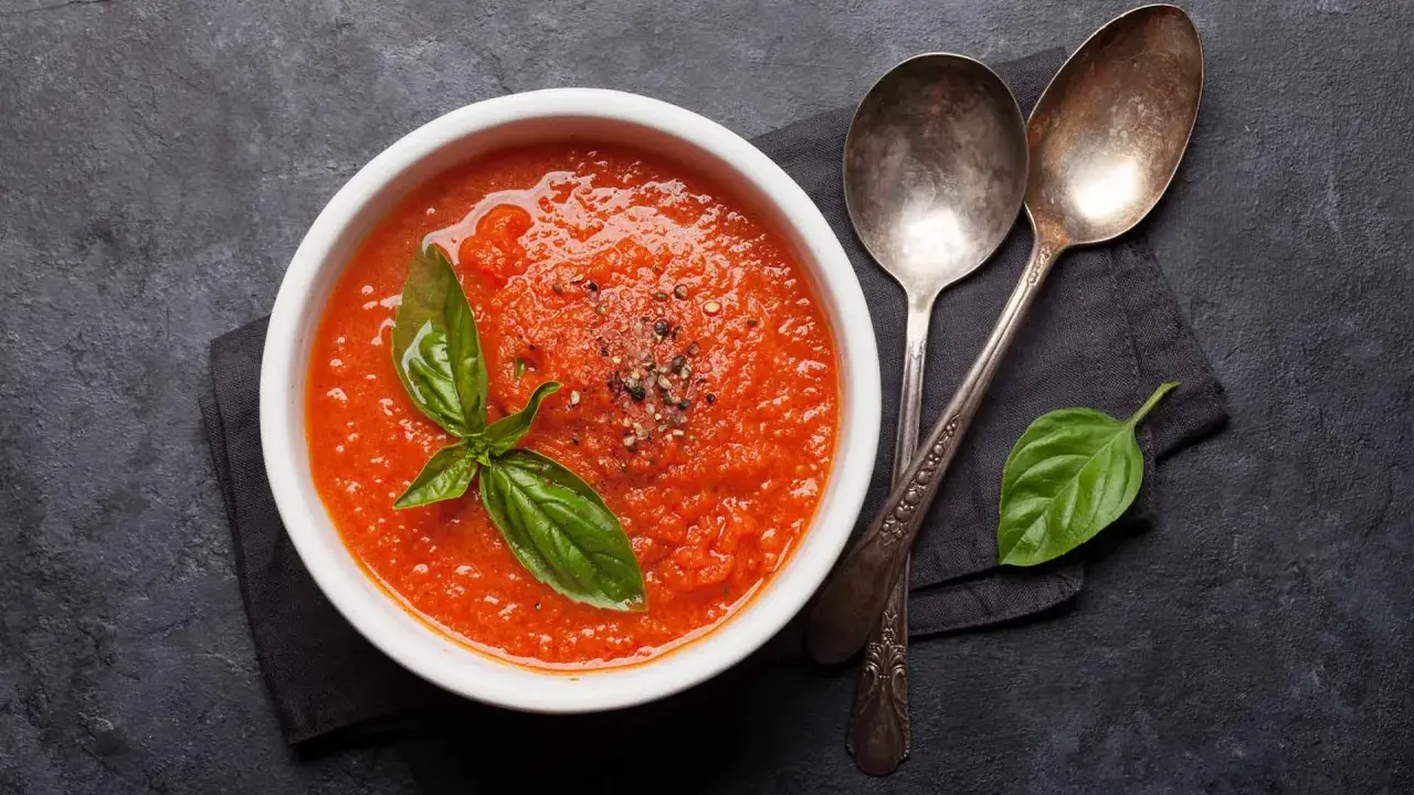 Schüssel mit selbstgemachter Tomatensauce, garniert mit Basilikum und Pfeffer, daneben zwei Löffel auf dunklem Hintergrund.