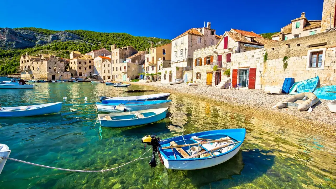 Barchette azzurre in un mare cristallino, un invito a scoprire dove andare al mare in Croazia. Case di pietra e colline verdi fanno da sfondo.