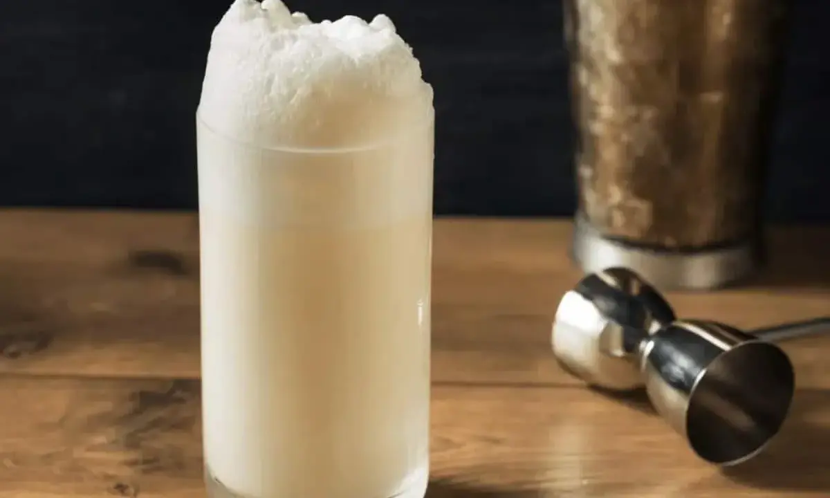 Ein cremiger Ramos Gin Fizz mit üppigem Schaum. Daneben ein Jigger und ein Shaker, bereit für den nächsten Drink.
