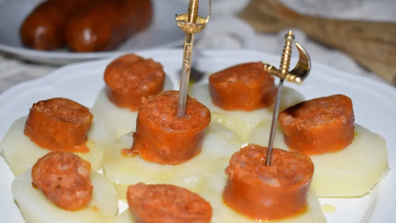 Chorizo a la sidra en rodajas, servido sobre patatas y adornado con espadas doradas.