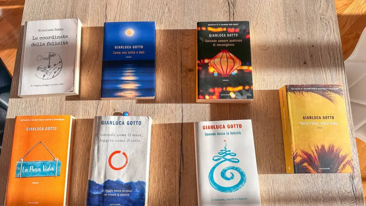 Una collezione di **libri** di Gianluca Gotto disposti su un tavolo di legno. Tra i titoli: "Le coordinate della felicità", "Come una notte a Bali", "La Pura Vida".
