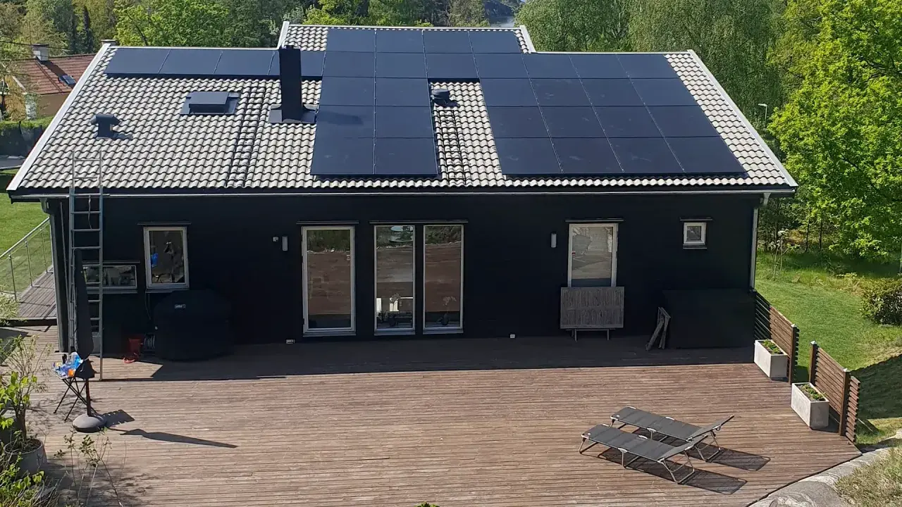 Ett mörkt hus med en liten solcellsanläggning på taket. En stor altan med solstolar leder ut mot en grönskande trädgård.