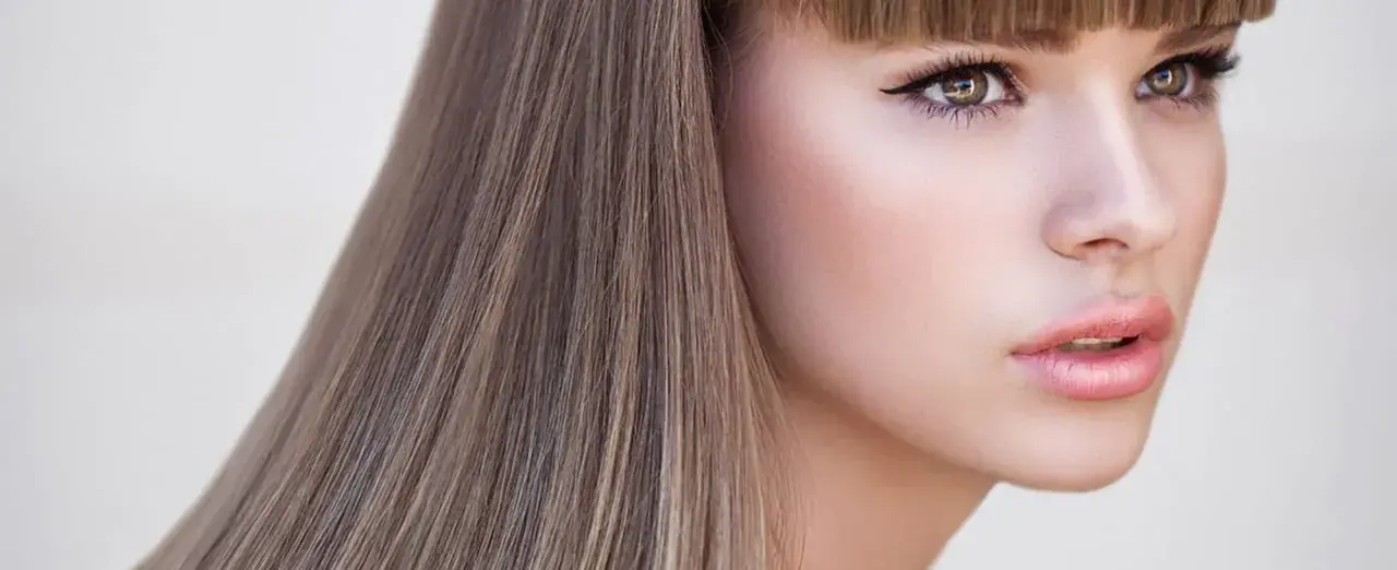 Mujer con cabello rubio champiñón, ojos delineados y labios rosados, con un fondo claro.