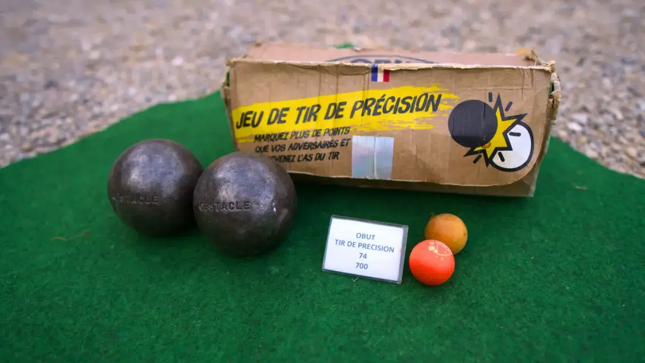 Deux boules de pétanque, un cochonnet et une boîte "Jeu de tir de précision" suggèrent un défi de poids et de diamètre pour le tireur.