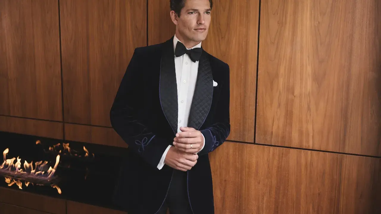 A man in a dark velvet tuxedo with a bow tie, embodying festive chic. He stands by a modern fireplace with flames.
