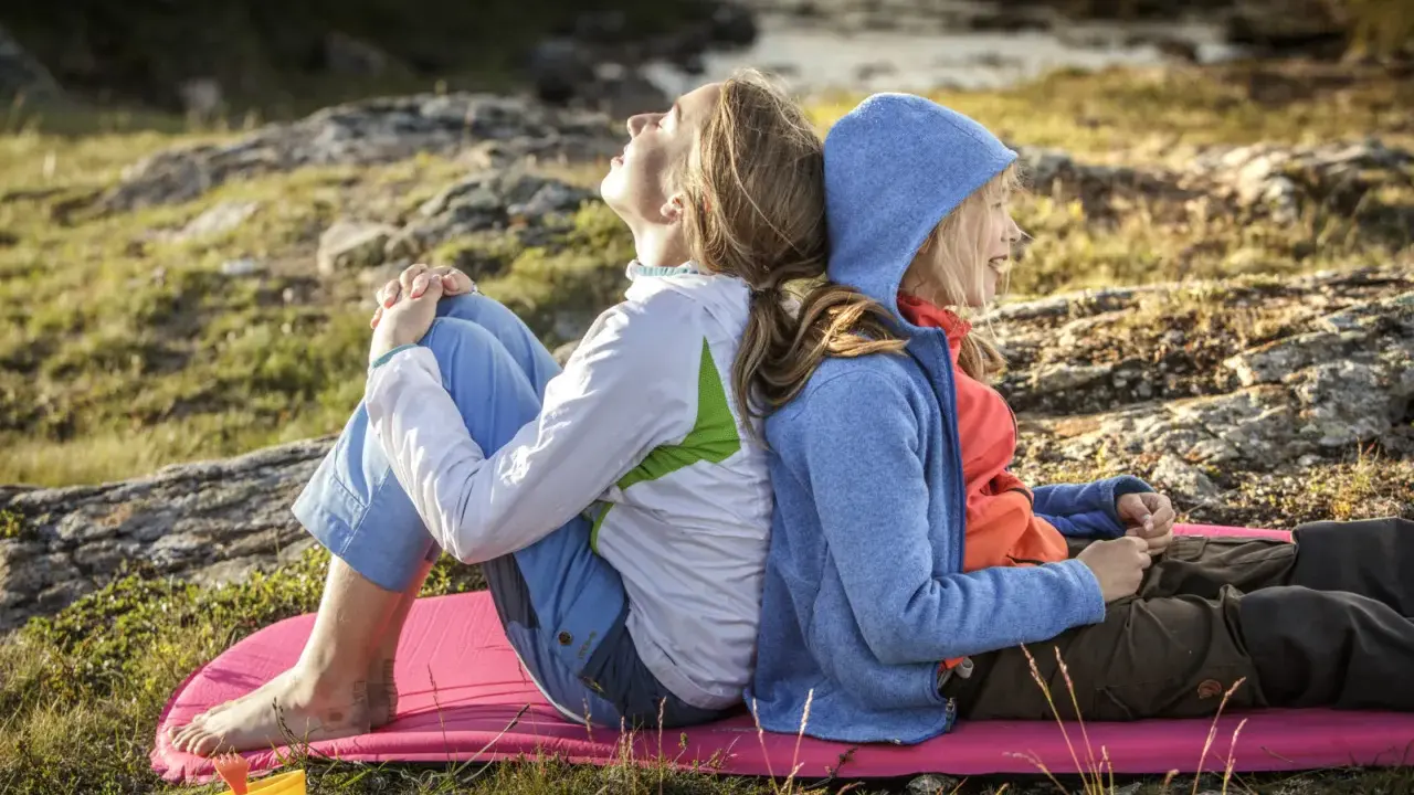Två personer, en vuxen och ett barn, sitter rygg mot rygg på en liggunderlag i naturen. En härlig stund att vandra med barn.