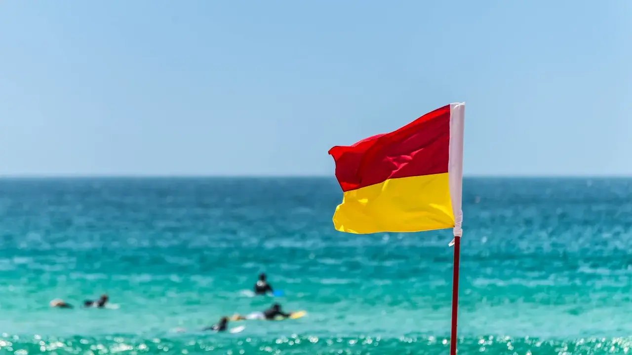 Gelb-rote Flagge am Strand weht im Wind. Surfer treiben auf dem türkisfarbenen Meer.