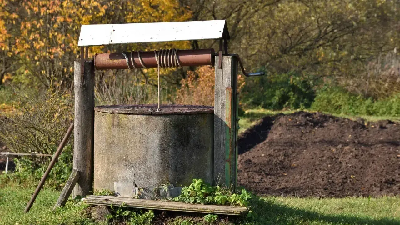 Stary, betonowy krąg studni kręgowej, z drewnianą konstrukcją i metalową korbą, otoczony jesienną zielenią.