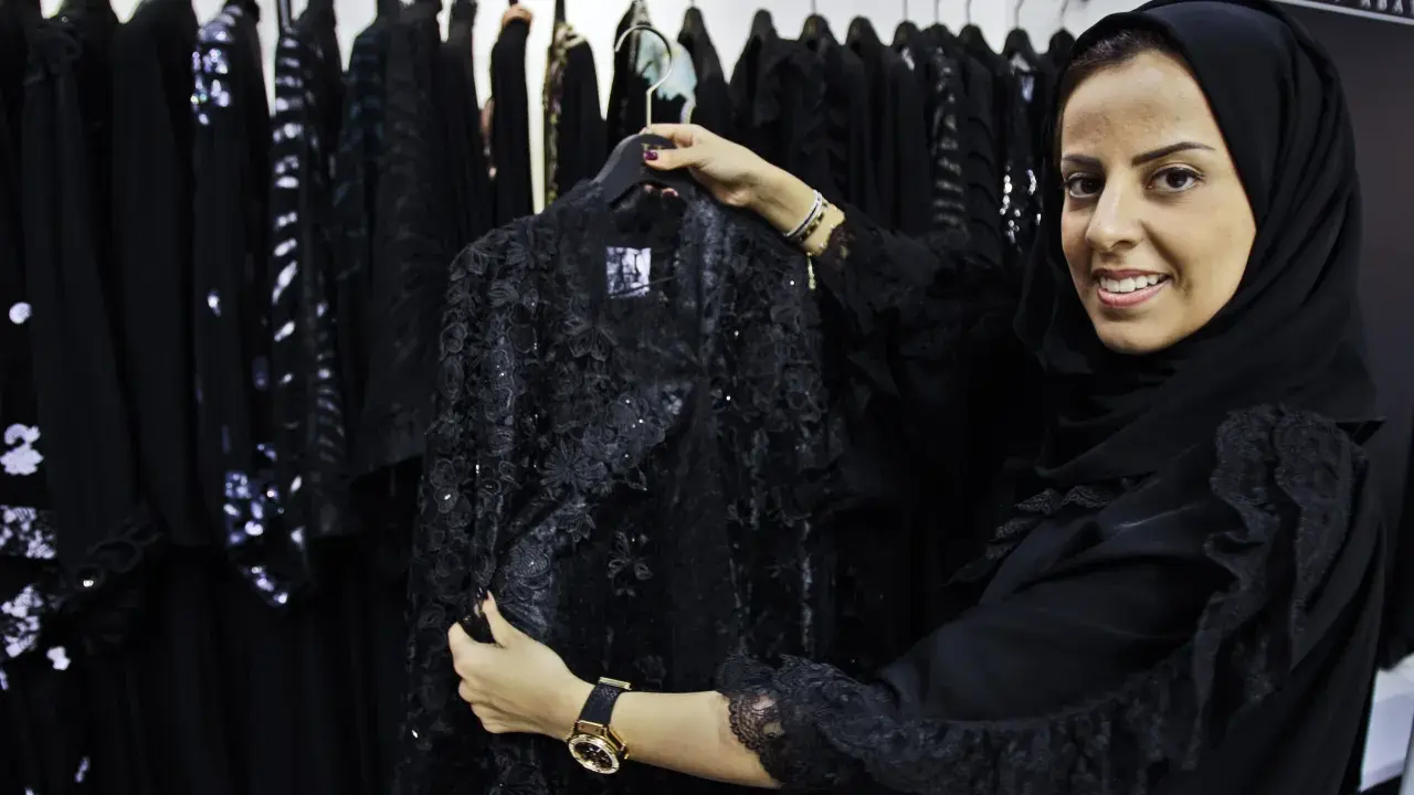 A woman smiles, holding a black lace abaya, showcasing elegant Saudi Arabia fashion.
