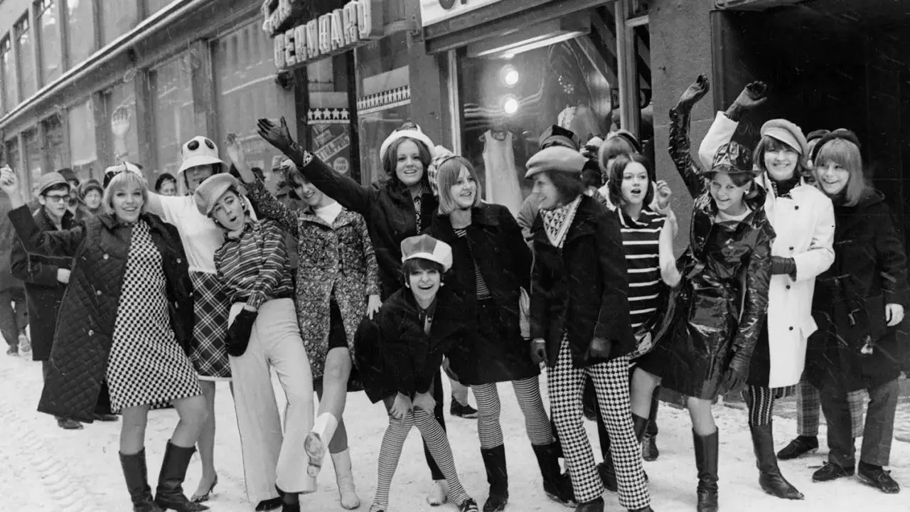 Ungdomar i trendiga kläder utanför en op-shop i Sverige 1960. Snö på marken och glada miner.