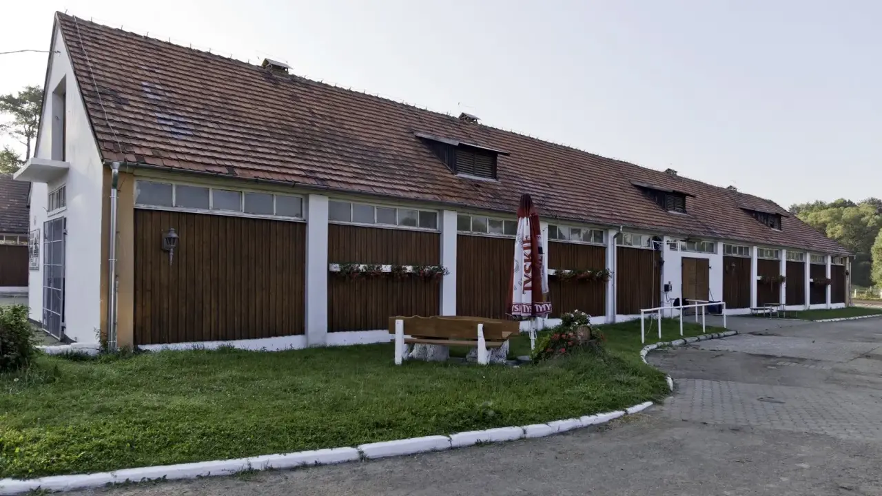Długi budynek stajni z drewnianymi drzwiami i oknami, z parasolem i ławką przed nim.