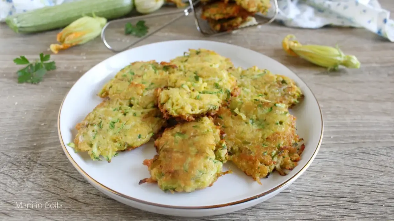 Buñuelos de calabacín dorados y crujientes en un plato blanco, con calabacines frescos y flores en el fondo.
