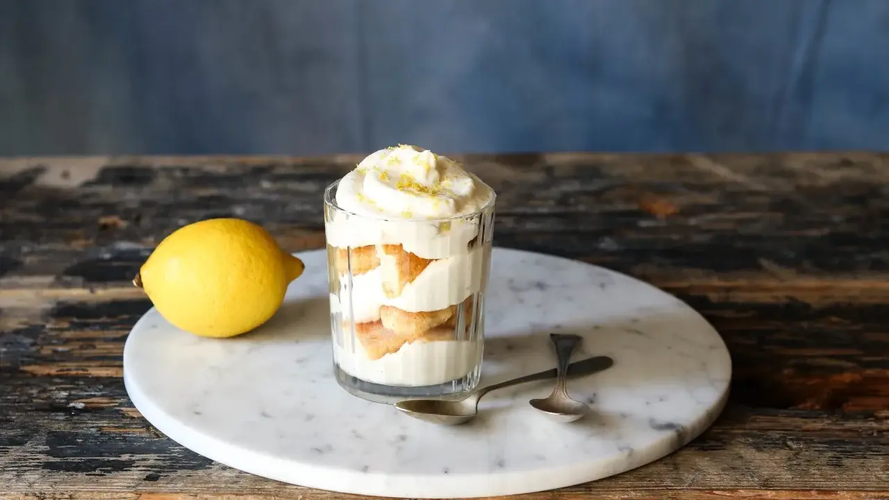 Ein Glas mit geschichtetem Zitronen Tiramisu, garniert mit Zitronenschale. Daneben liegt eine Zitrone.