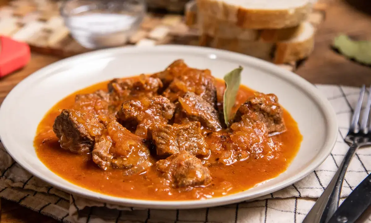 Sabrosa carne con tomate, guisada lentamente hasta quedar tierna, servida en un plato blanco con pan y sal al lado.
