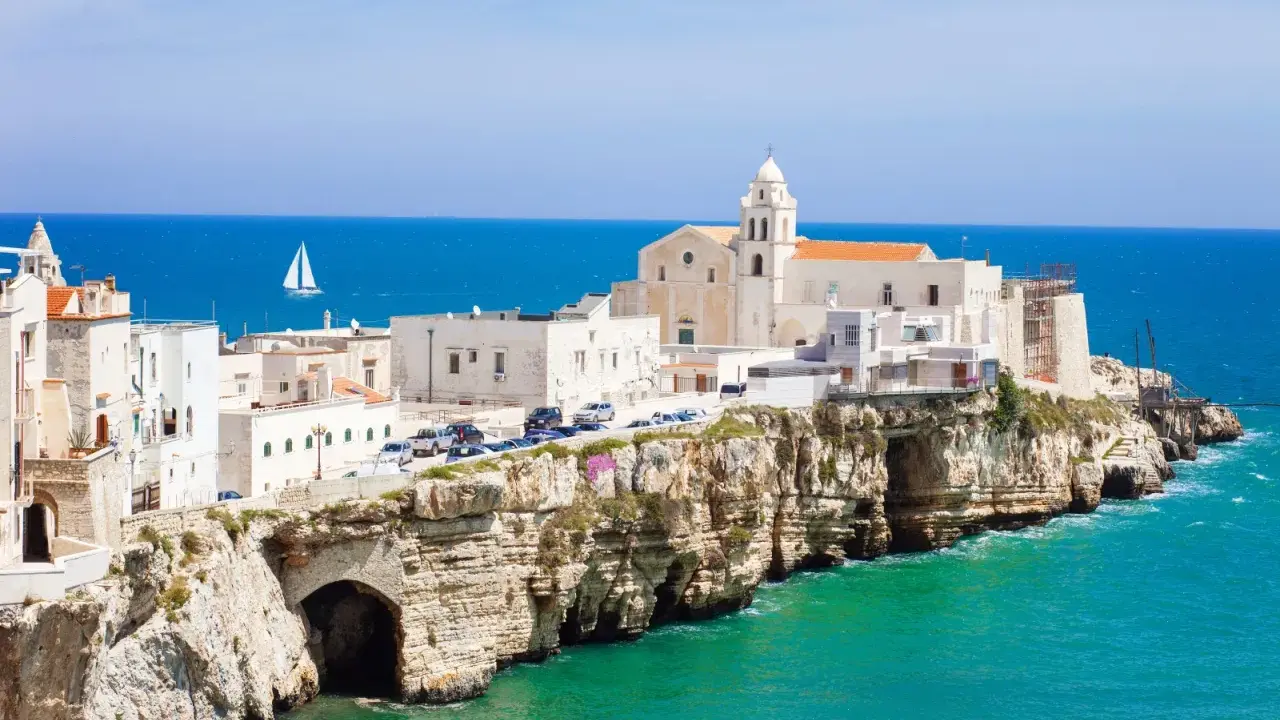 Un borgo bianco su scogliere a picco sul mare turchese, con una chiesa imponente. Un assaggio di cosa vedere in Puglia, con un veliero all'orizzonte.