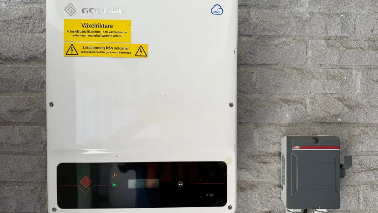 En vit GoodWe växelriktare 10kW monterad på en grå stenvägg. Bredvid sitter en grå ABB-box.