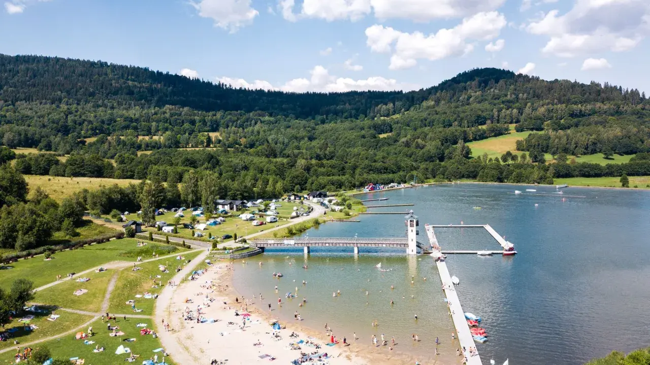 Słoneczny dzień nad zalewem w Starej Morawie. Ludzie odpoczywają na plaży i w wodzie, a w tle widać zielone wzgórza.