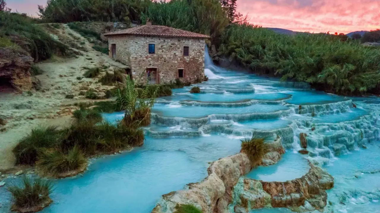 Cascate termali turchesi in Toscana, con un antico mulino in pietra e un cielo rosa al tramonto. Curiosità sulla Toscana: un luogo magico.