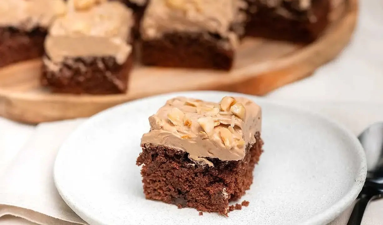 En bit chokladtårta med krämig frosting och nötter. Flera bitar serveras på ett träfat i bakgrunden.