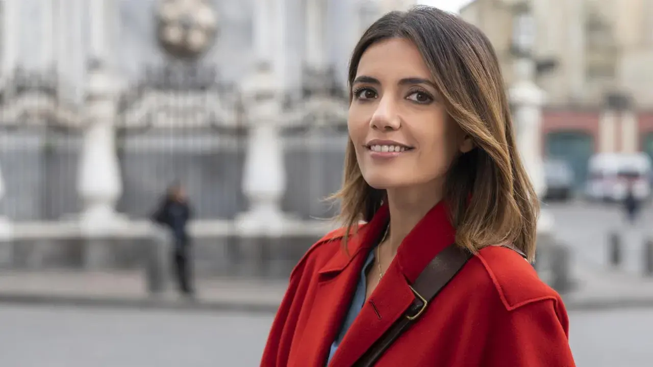 Donna con cappotto rosso sorride, con uno sfondo sfocato di edifici e una persona. Forse sta pensando ai **mina settembre libri** da leggere.