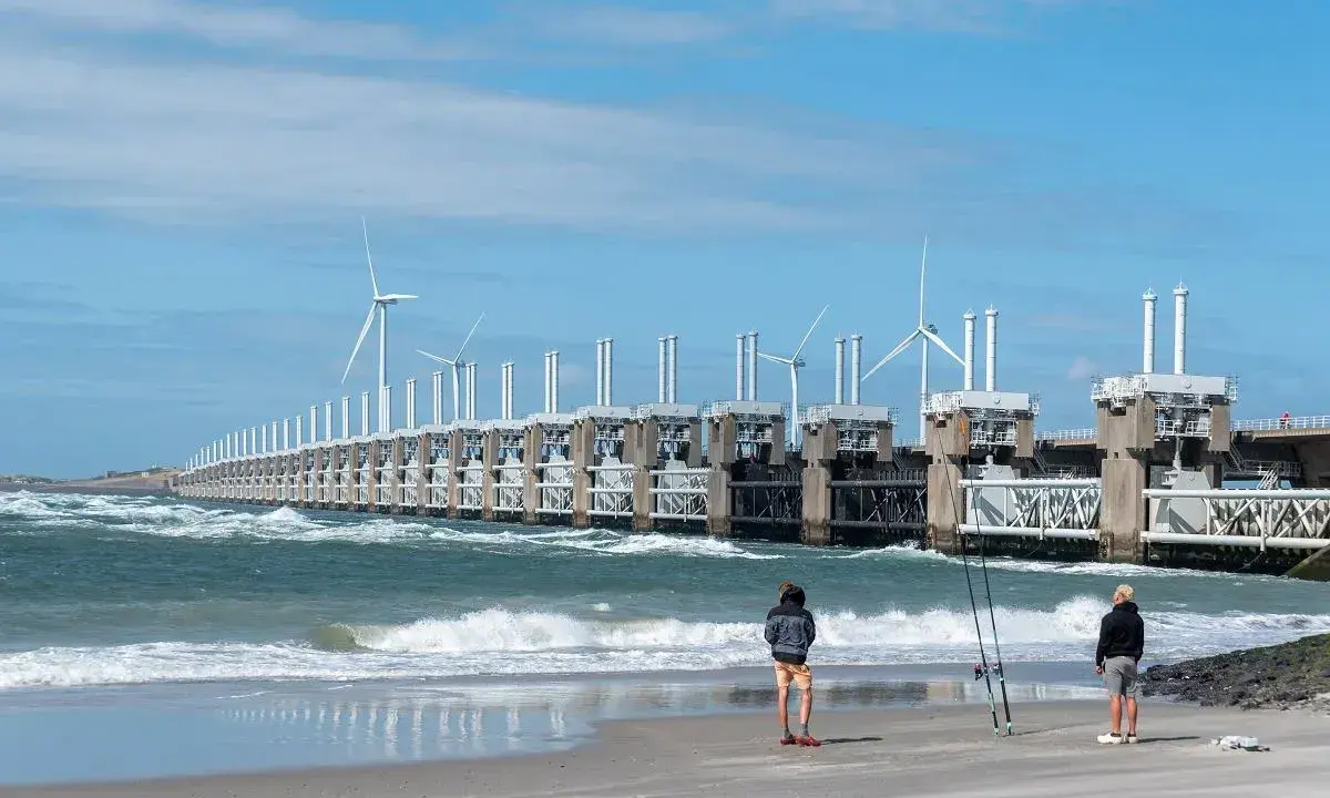 Dwie osoby stoją na plaży, obserwując falujące morze i imponującą zaporę z turbinami wiatrowymi. Takie morze jest w Holandii, chronione przez innowacyjne konstrukcje.