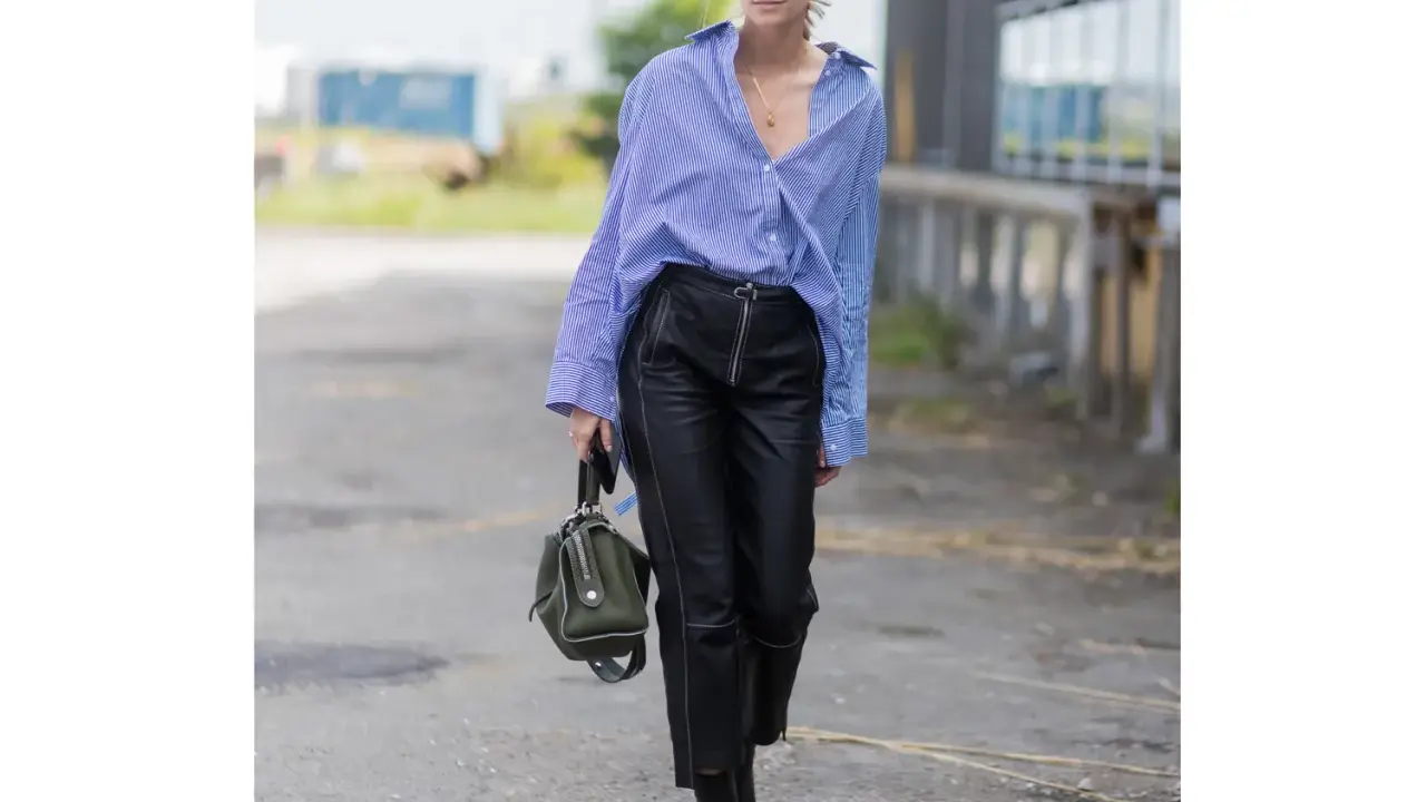Donna con camicia a righe blu e bianche, occhiali da sole e un elegante outfit pantaloni ecopelle neri con zip frontale. Porta una borsa verde oliva.