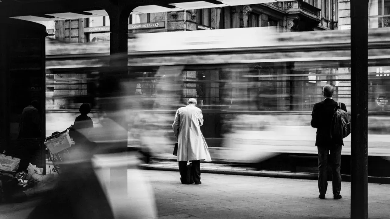 Street photography esempi: un tram sfreccia, lasciando figure sfocate in attesa alla fermata.
