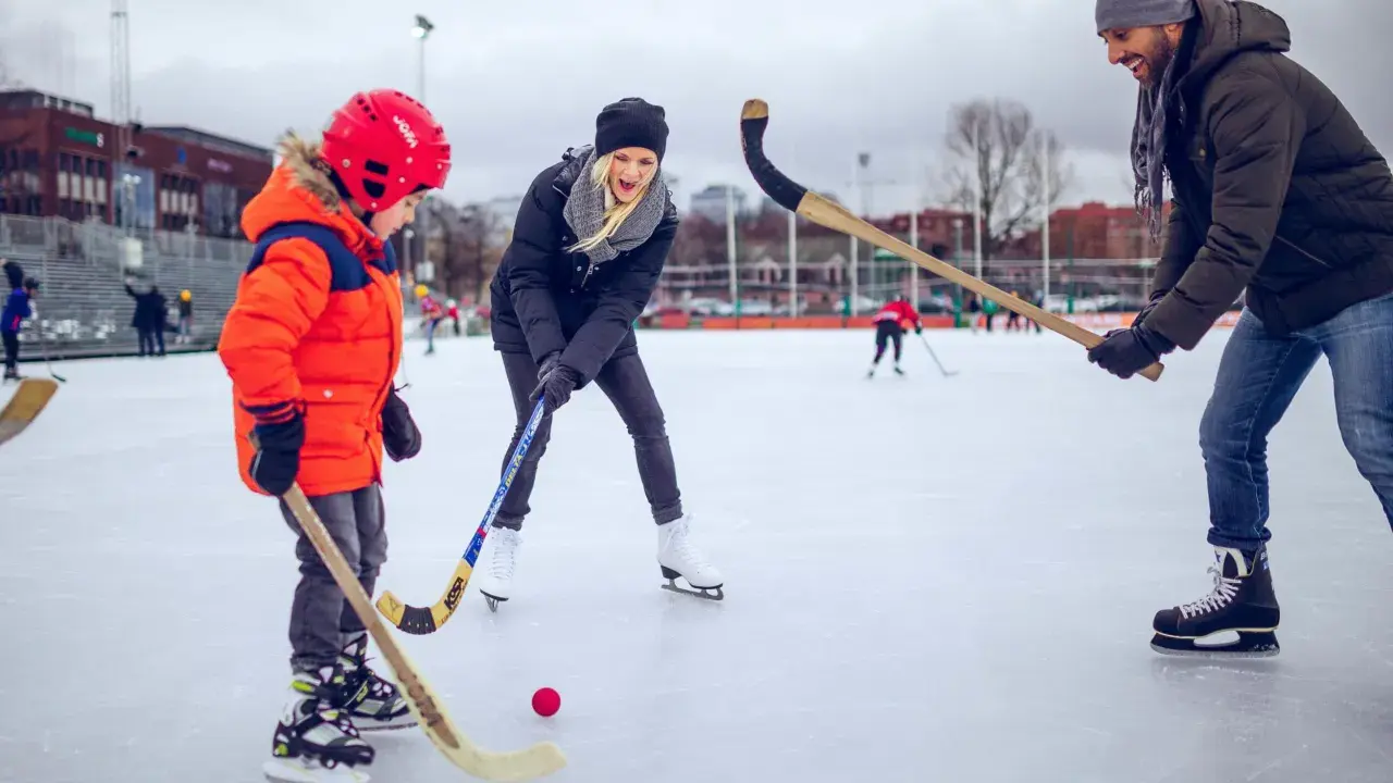 Familj njuter av ishockey i Göteborg. En pappa, mamma och son med hockeyklubbor och en röd puck på isen.