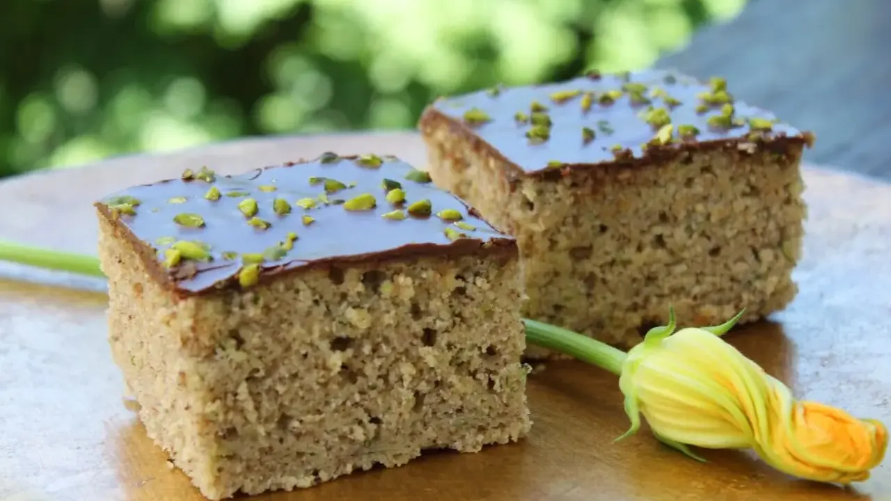 Zwei Stücke saftiger Zucchini Kuchen mit Schokoladenglasur und gehackten Pistazien, daneben eine Zucchiniblüte.